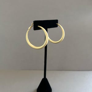 May include: A pair of gold-colored hoop earrings displayed on a black stand. The earrings are circular with a smooth, matte finish. The background is a solid, neutral gray.