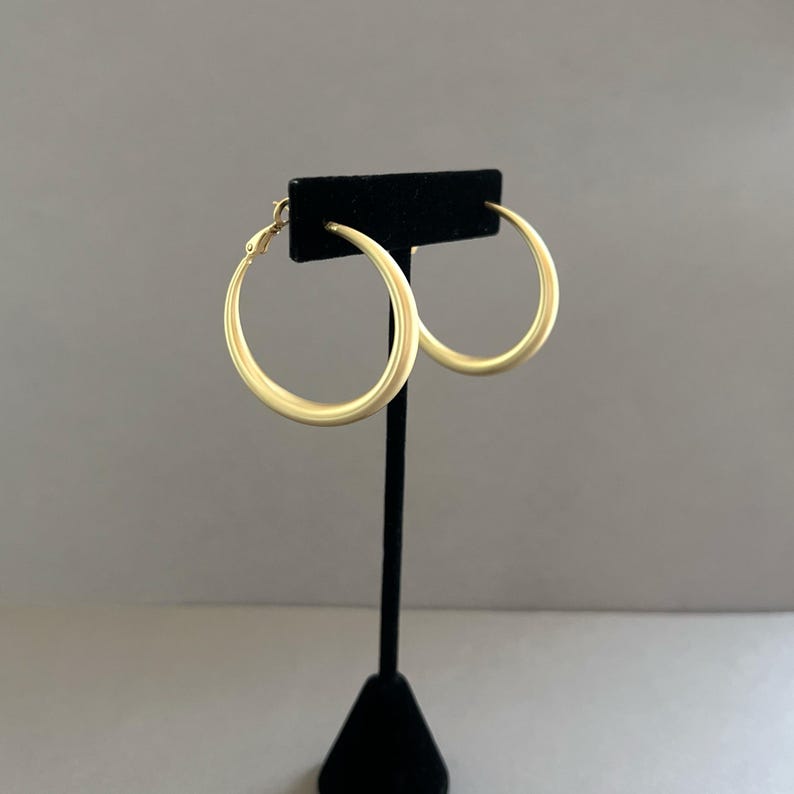 May include: A pair of gold-colored hoop earrings displayed on a black stand. The earrings are circular with a smooth, matte finish. The background is a solid, neutral gray.