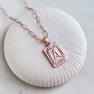 Könnte beinhalten: Halskette in Roségold mit einem rechteckigen Anhänger mit dem Buchstaben "A". Der Anhänger hat einen strukturierten Rand und hängt an einer filigranen Kette. Die Halskette wird auf einer weißen, strukturierten Oberfläche präsentiert.