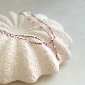 Puede incluir: Un delicado collar de oro rosa con un pequeño colgante de ópalo ovalado. El collar presenta un diseño de cadena de doble hebra y se muestra sobre una superficie texturizada blanquecina. El colgante es de color blanco lechoso.