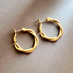 Puede incluir: Un par de pendientes de aro dorados. Cada pendiente presenta un diseño retorcido, creando una apariencia texturizada. Los pendientes se muestran sobre una superficie gris neutra, resaltando su brillo metálico. Los pendientes tienen un estilo clásico y elegante.