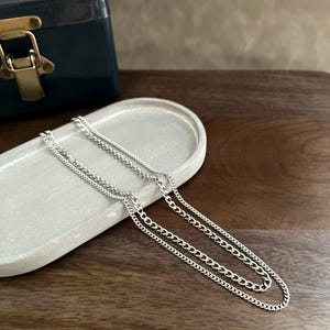 May include: Silver chain necklaces displayed on a gray oval tray. The necklaces have different chain styles, including a curb chain. A dark teal box with a gold clasp is in the background. The tray is on a dark wood surface.
