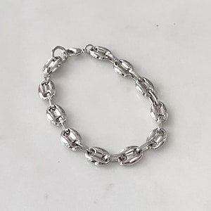 May include: A silver-colored bracelet with a coffee bean link design. The bracelet is made of a shiny metal and has a lobster clasp closure. The bracelet is shown on a white surface.