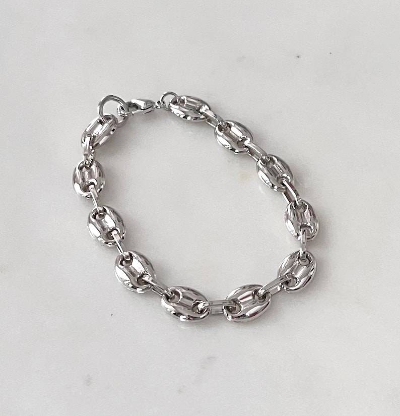 May include: A silver-colored bracelet with a coffee bean link design. The bracelet is made of a shiny metal and has a lobster clasp closure. The bracelet is shown on a white surface.