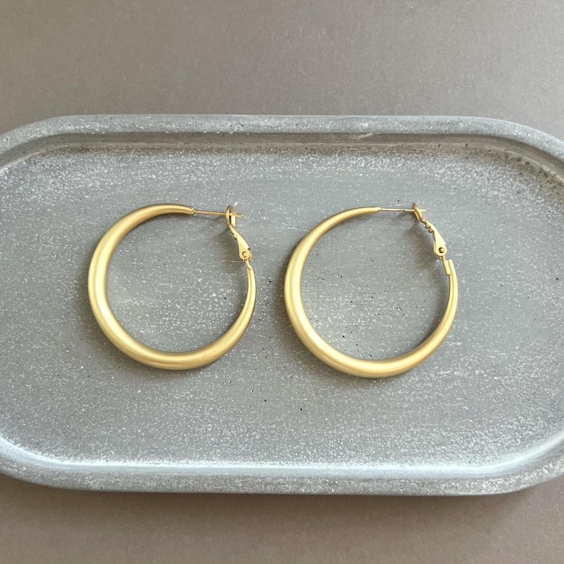 May include: A pair of gold-colored hoop earrings. The earrings are round and have a matte finish. They are displayed on a gray, oval-shaped tray. The earrings are a classic jewelry accessory.