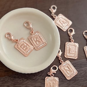 May include: Rose gold letter charms with lobster clasps. The charms are rectangular with a decorative border and feature the letters A, B, K, L, M, and N. The charms are displayed in a white dish and on a dark wooden surface.
