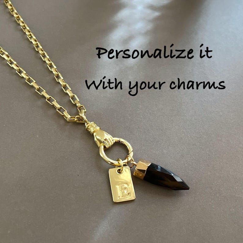 May include: A gold-toned chain necklace featuring a hand-shaped clasp holding a circular ring. Attached to the ring are a rectangular gold tag engraved with the letter E and a dark, pointed stone pendant. Text reads: Personalize it with your charms.