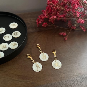 May include: Three gold-coloured initial charms with white, iridescent circular pendants, each featuring a different letter. Additional initial pendants are in a black tray. Dried pink flowers are in the background.
