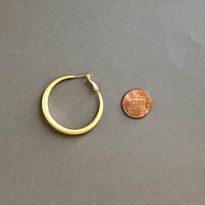 May include: A gold-colored hoop earring with a simple design. The earring is next to a penny for size comparison. The earring has a clasp closure. The background is a solid gray color.