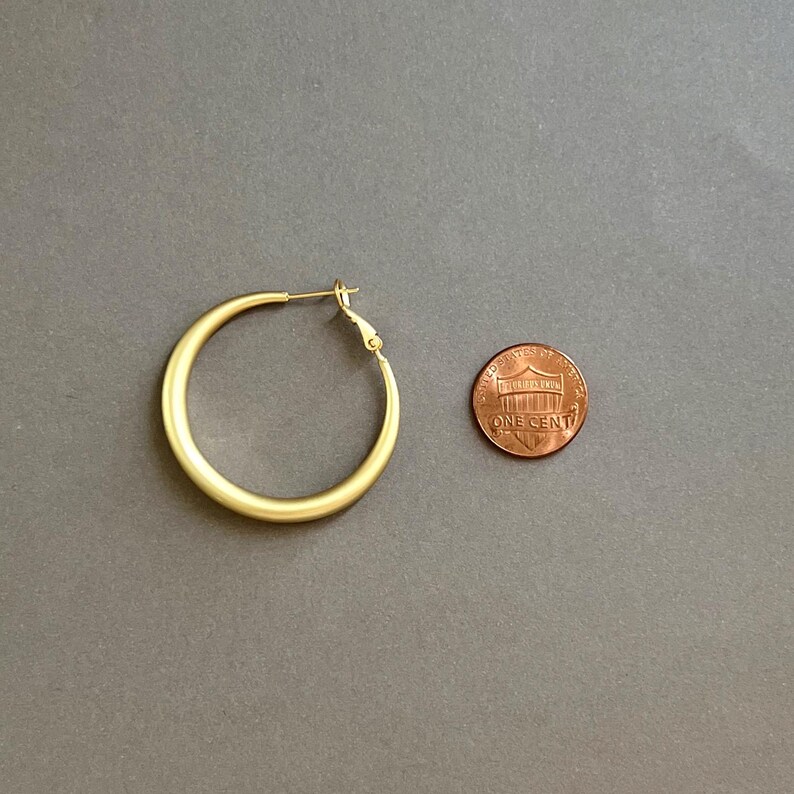 May include: A gold-colored hoop earring with a simple design. The earring is next to a penny for size comparison. The earring has a clasp closure. The background is a solid gray color.