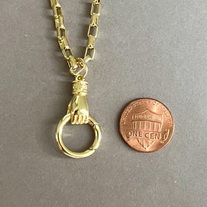 May include: A gold-toned necklace featuring a chain with rectangular links and a pendant shaped like a hand holding a circular ring. The pendant is positioned next to a United States one-cent coin for scale, which has a diameter of 0.75 inches.