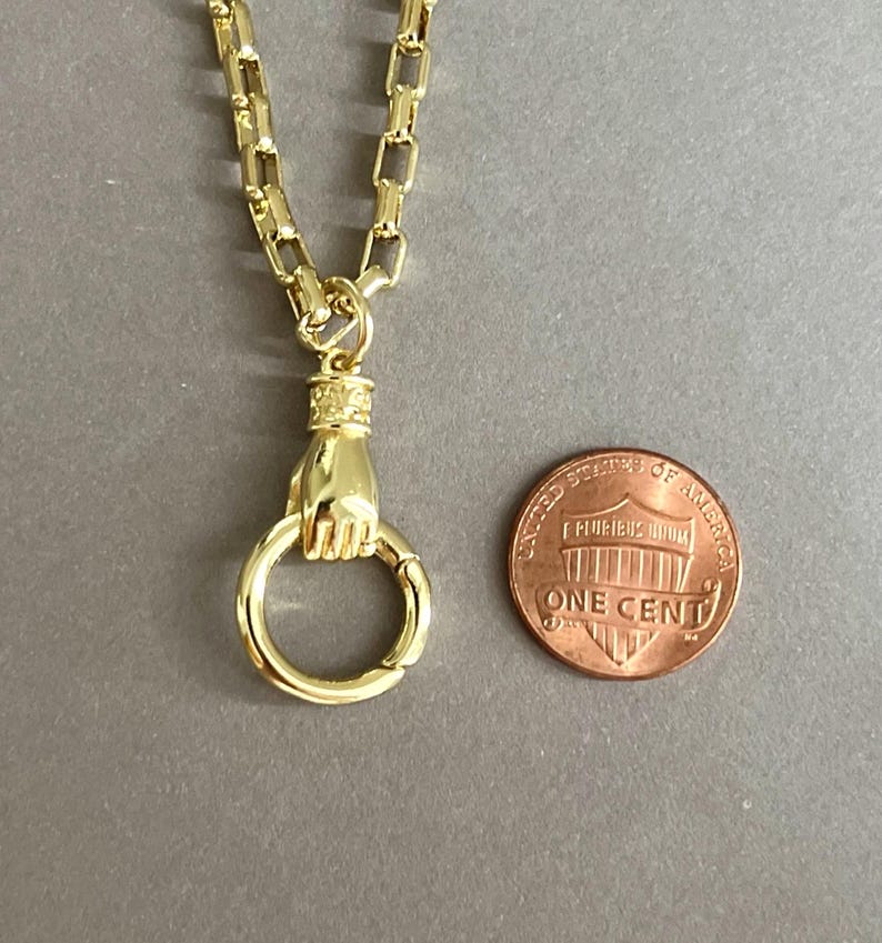 May include: A gold-toned necklace featuring a chain with rectangular links and a pendant shaped like a hand holding a circular ring. The pendant is positioned next to a United States one-cent coin for scale, which has a diameter of 0.75 inches.