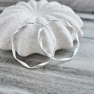 May include: A pair of silver-toned hoop earrings with a twisted design. The earrings have a matte finish and a simple clasp closure. The earrings are displayed on a white, textured surface against a marble background. Jewelry.