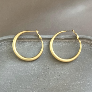 May include: A pair of gold-colored hoop earrings. The earrings are circular with a simple, elegant design. The hoops are displayed on a gray surface. The earrings have a matte finish and a classic style.