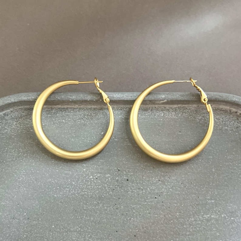May include: A pair of gold-colored hoop earrings. The earrings are circular with a simple, elegant design. The hoops are displayed on a gray surface. The earrings have a matte finish and a classic style.