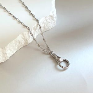 May include: A silver-toned necklace featuring a delicate chain with textured links and a pendant shaped like a hand holding a circular ring. The metal has a polished finish, and the design includes a small cuff detail at the wrist of the hand.