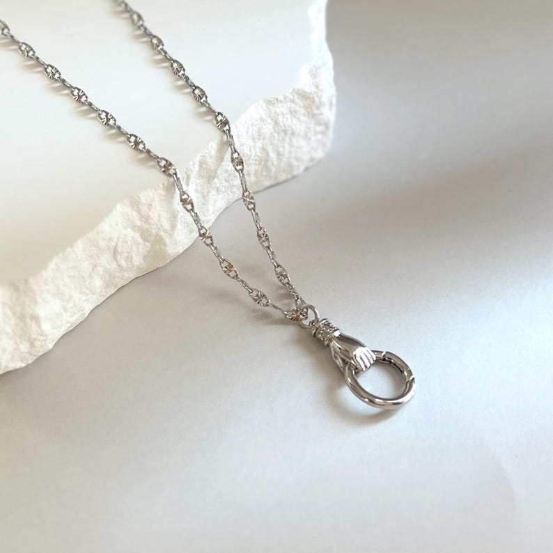 May include: A silver-toned necklace featuring a delicate chain with textured links and a pendant shaped like a hand holding a circular ring. The metal has a polished finish, and the design includes a small cuff detail at the wrist of the hand.