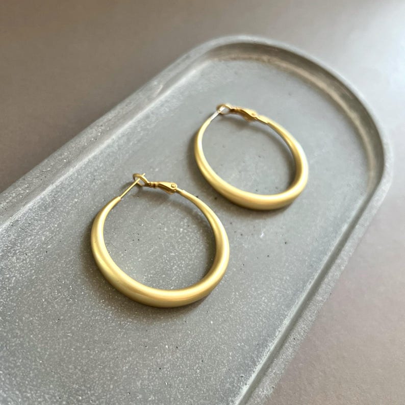 May include: Two gold-colored hoop earrings with a matte finish are displayed on a gray, rectangular tray. The earrings have a classic, rounded design and a simple clasp closure. The background is a neutral gray.