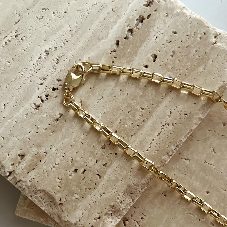 May include: A gold-toned chain necklace featuring a series of interconnected square-shaped links. The chain includes a small, circular lobster claw clasp mechanism at the end, which is securely attached to the final link of the metallic jewelry piece.