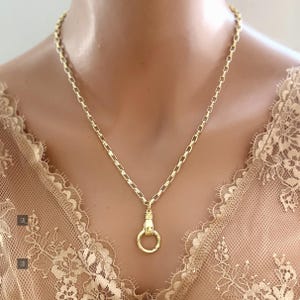May include: A gold-toned necklace featuring a chain with rectangular links and a pendant shaped like a hand holding a circular ring. The metal has a polished finish, and the design is centered on a delicate, light-colored lace garment.