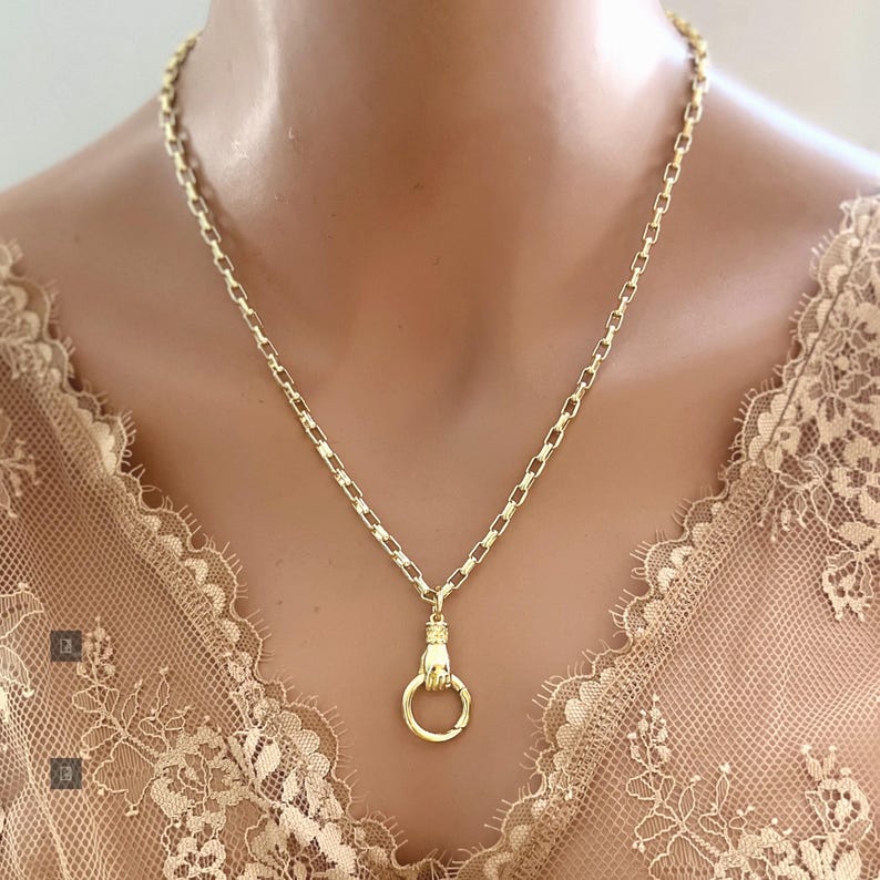 May include: A gold-toned necklace featuring a chain with rectangular links and a pendant shaped like a hand holding a circular ring. The metal has a polished finish, and the design is centered on a delicate, light-colored lace garment.