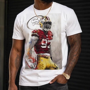 May include: A white short-sleeved t-shirt featuring a graphic print of a football player in a red and gold uniform with the number 95. The design includes a faded 49ers logo in the background, rendered in a watercolor style with paint splatters.