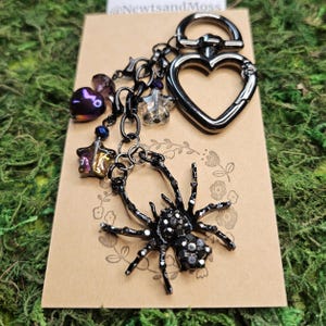 A black spider charm keychain with a heart-shaped clasp. Rhinestone-studded Spider. The keychain also features purple heart and star glass charms.