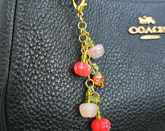 May include: A gold-toned metal charm featuring a lobster clasp and a chain adorned with small green glass beads, translucent pink and orange fruit-shaped beads, and green leaf-shaped accents, attached to a black textured leather handbag.
