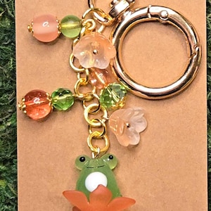 May include: A gold-toned metal keychain featuring a green glass frog charm sitting on an orange flower. The accessory includes translucent pink and green beads, small flower-shaped charms, and a circular gold ring clasp attached to a gold chain.