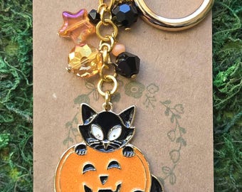 May include: A gold-toned metal keychain featuring a black cat enamel charm emerging from an orange jack-o'-lantern. The keychain includes a gold ring and a decorative chain adorned with faceted black, orange, and iridescent star-shaped beads.