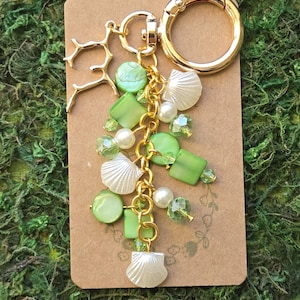 May include: A gold-toned keychain featuring a collection of green and white beads, small white seashell charms, and a gold-toned coral-shaped charm. The keychain is attached to a gold-toned circular ring and is displayed on a rectangular brown card.