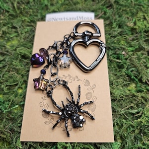 K&ouml;nnte beinhalten: Ein herzf&ouml;rmiger Schl&uuml;sselanh&auml;nger aus schwarzem Metall, befestigt an einer Kette mit einem schwarzen Spinnenanh&auml;nger, einer violetten Herzperle und zwei sternf&ouml;rmigen Perlen. Der Anh&auml;nger ist auf einer beigen Karte mit dem Text @NewtsandMoss.