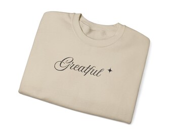 May include: A beige sweatshirt with the word "Greatful" in a cursive font, accompanied by a small star. The sweatshirt is folded neatly, showcasing the design on the front. The overall aesthetic is simple and minimalist.