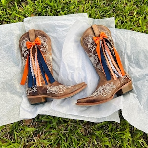 K&ouml;nnte beinhalten: Ein Paar braune Leder-Cowboystiefel mit floraler Stickerei und spitzer Zehenpartie. Jeder Stiefel ist mit einer orangefarbenen Schleife und farbigen B&auml;ndern in Orange, Wei&szlig; und Marineblau verziert. Die Stiefel werden auf einer wei&szlig;en Oberfl&auml;che vor gr&uuml;nem Gras pr&auml;sentiert.