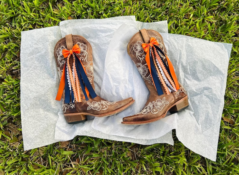 K&ouml;nnte beinhalten: Ein Paar braune Leder-Cowboystiefel mit floraler Stickerei und spitzer Zehenpartie. Jeder Stiefel ist mit einer orangefarbenen Schleife und farbigen B&auml;ndern in Orange, Wei&szlig; und Marineblau verziert. Die Stiefel werden auf einer wei&szlig;en Oberfl&auml;che vor gr&uuml;nem Gras pr&auml;sentiert.