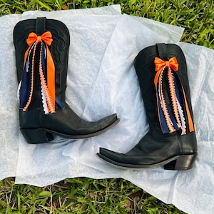 K&ouml;nnte beinhalten: Ein Paar schwarze Cowboy-Stiefel mit spitzer Zehenpartie, verziert mit orangefarbenen Schleifen und passenden B&auml;ndern. Die B&auml;nder sind mit marineblauer und wei&szlig;er Spitze akzentuiert, was einen festlichen Look erzeugt. Die Stiefel liegen auf einer wei&szlig;en Oberfl&auml;che.