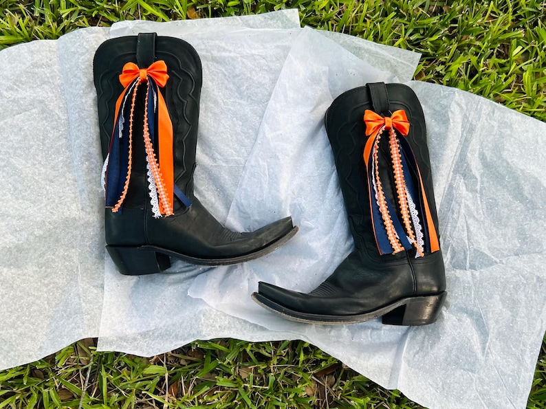 K&ouml;nnte beinhalten: Ein Paar schwarze Cowboy-Stiefel mit spitzer Zehenpartie, verziert mit orangefarbenen Schleifen und passenden B&auml;ndern. Die B&auml;nder sind mit marineblauer und wei&szlig;er Spitze akzentuiert, was einen festlichen Look erzeugt. Die Stiefel liegen auf einer wei&szlig;en Oberfl&auml;che.