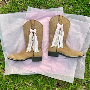 Puede incluir: Un par de botas vaqueras de ante color tostado con un patr&oacute;n floral en relieve en la ca&ntilde;a y tac&oacute;n negro. Cada bota est&aacute; adornada con un lazo de cinta rosa claro y adornos colgantes de encaje blanco y cuentas tipo perla.