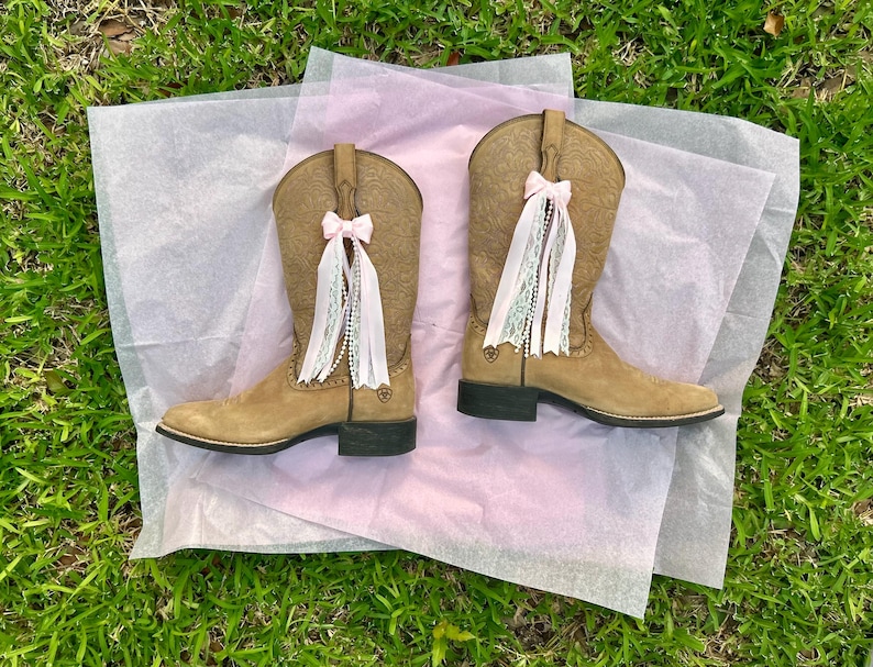 Puede incluir: Un par de botas vaqueras de ante color tostado con un patr&oacute;n floral en relieve en la ca&ntilde;a y tac&oacute;n negro. Cada bota est&aacute; adornada con un lazo de cinta rosa claro y adornos colgantes de encaje blanco y cuentas tipo perla.