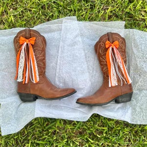 May include: A pair of brown leather cowboy boots with decorative orange bows and hanging ribbons. The ribbons include white lace, orange fabric, and strings of white beads. The boots are on a sheet of white paper, set against a backdrop of green grass.