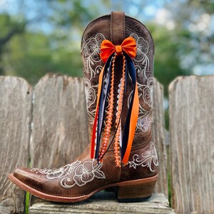 K&ouml;nnte beinhalten: Brauner Leder-Cowboystiefel mit floraler Stickerei und einer orangefarbenen Schleife. Der Stiefel hat eine Mandelspitze und einen gestapelten Absatz. Orange, marineblaue und wei&szlig;e B&auml;nder und Perlenstr&auml;nge sind am Stiefel befestigt.