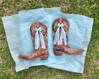 Op de afbeelding: Een paar bruine leren cowboylaarzen met witte bloemenborduursels. Elke laars is versierd met een lichtblauwe satijnen strik, witte kanten rand en een streng witte kunstparels bevestigd aan de voorzijde van de schacht.