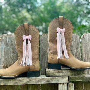 Puede incluir: Un par de botas vaqueras de cuero color canela con un patr&oacute;n floral en relieve en la ca&ntilde;a y tac&oacute;n negro. Cada bota est&aacute; adornada con un lazo de cinta rosa claro con encaje y adornos de cuentas tipo perla que cuelgan del lateral.