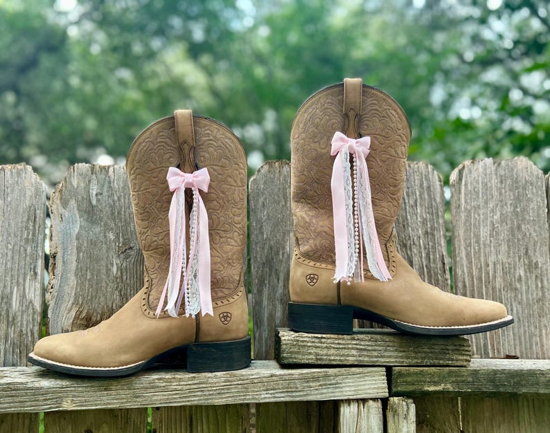 Puede incluir: Un par de botas vaqueras de cuero color canela con un patr&oacute;n floral en relieve en la ca&ntilde;a y tac&oacute;n negro. Cada bota est&aacute; adornada con un lazo de cinta rosa claro con encaje y adornos de cuentas tipo perla que cuelgan del lateral.