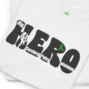 Puede incluir: Dos camisetas blancas apiladas. La camiseta superior muestra el texto "My HERO Cerebral Palsy WARRIOR" en blanco y negro, con una cinta verde. Las camisetas están dobladas.