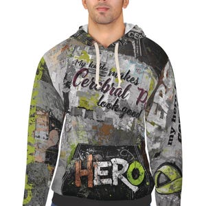 Op de afbeelding: Een hoodie met een kleurrijk, abstract ontwerp. De voorkant toont de tekst "My kiddo makes Cerebral Palsy look good" in een cursief lettertype, met het woord "HERO" in vette letters eronder. Het ontwerp bevat tinten grijs, groen en oranje.