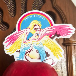 Puede incluir: Una pegatina con una ilustraci&oacute;n estilizada de una figura alada con cabello amarillo y alas coloridas. Las alas son rosas, amarillas y azules. El texto "PAN KING" est&aacute; en un c&iacute;rculo azul.