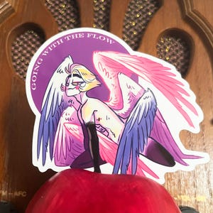 Puede incluir: Una pegatina con una ilustraci&oacute;n estilizada de una figura alada con cabello rubio, alas rosas y azules, y el texto "GOING WITH THE FLOW" en un c&iacute;rculo morado. La figura est&aacute; arrodillada sobre una superficie roja.