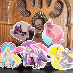 May include: A collection of colorful stickers featuring stylized illustrations of figures with wings and animalistic features. The stickers have text that reads "ACE IN THE HOLE", "PAN KING", and "LOGICALLY ACCURATE". The stickers are placed on a vintage wooden radio.