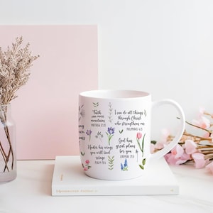 Może przedstawiać: Biały ceramiczny kubek ozdobiony kwiatowymi ilustracjami i inspirującymi cytatami. Kubek ma zakrzywiony uchwyt i stoi na białej książce. Cytaty to "Wiara może góry przenosić" i "Wszystko mogę w Tym, który mnie umacnia".